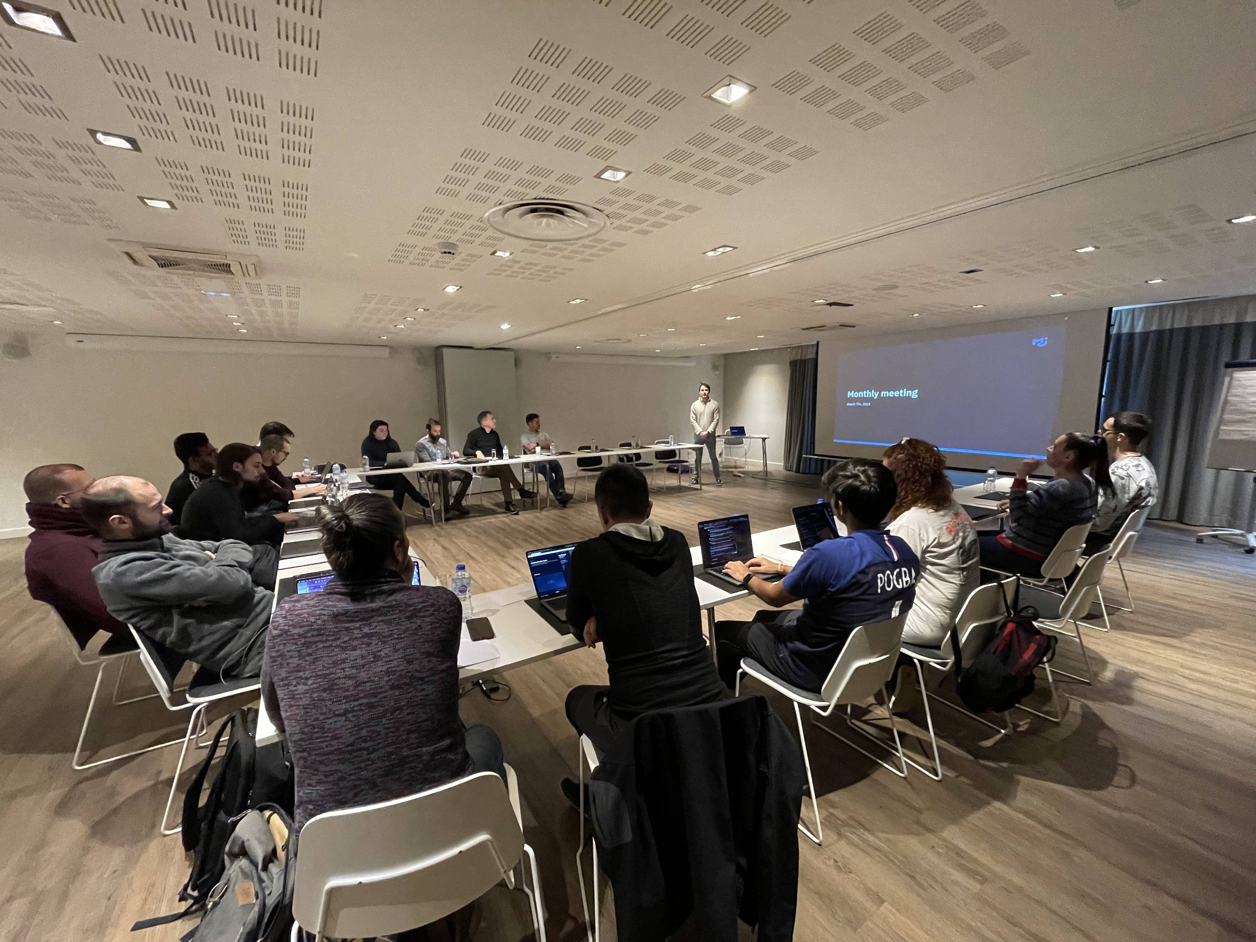 MUI team members sit in a half-U shape as Co-Founder Olivier Tassinari presents the monthly meeting slides.