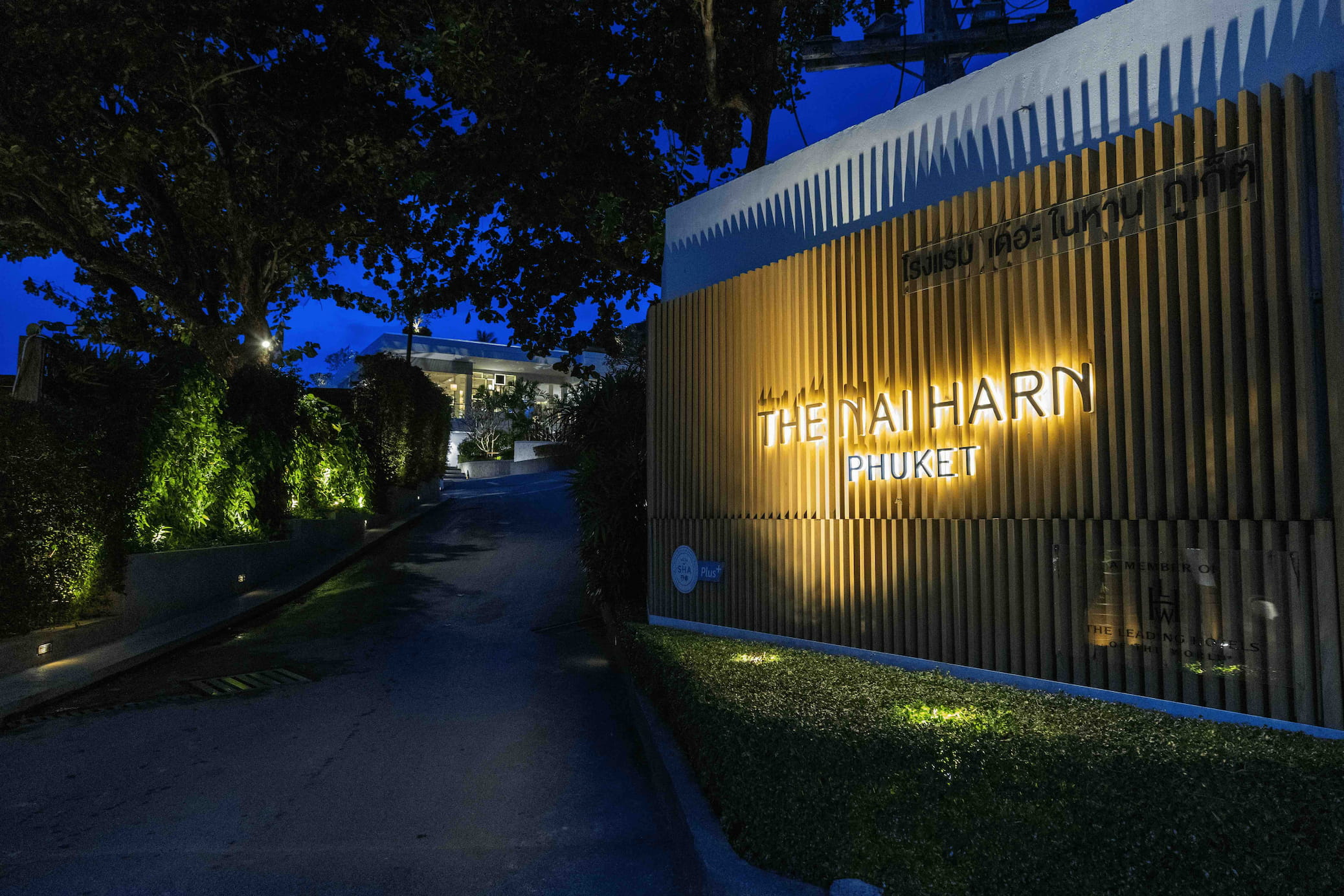Entrance to the Nai Harn property at night. A lit sign on a leafy backdrop reads: The Nai Harn - Phuket.
